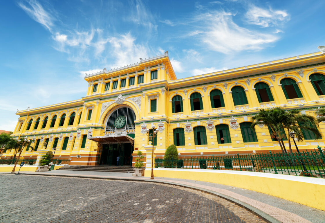 post office in ho chi minh, vietnam, a long yellow building with arched windows all along with green shutters and in the centre there is a wider and taller part of the building with a large clock in the middle and wide open doors after a few steps
