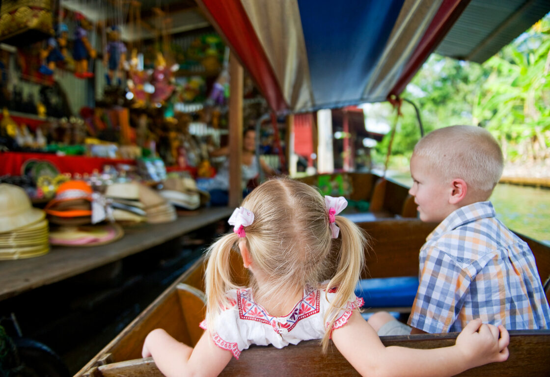 a young girl and boy sit in a wooden boat next to a stall with hats and other items at a floating market in thailand