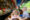 a young girl and boy sit in a wooden boat next to a stall with hats and other items at a floating market in thailand