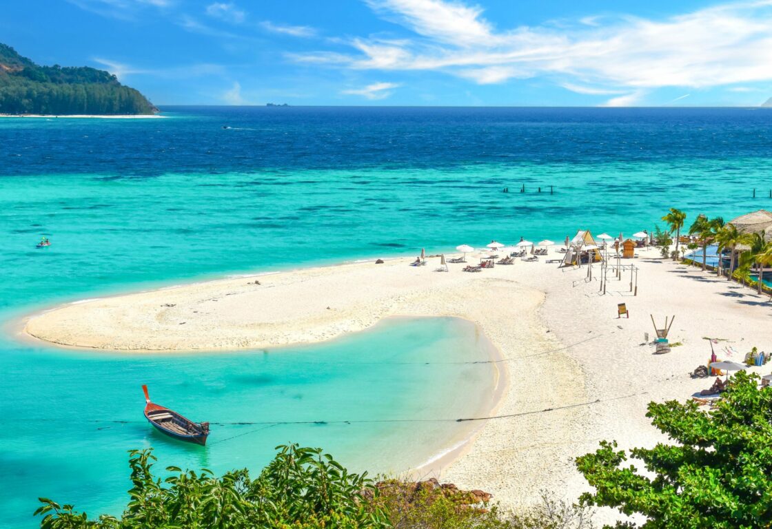 a sea with different shades of blue surround a piece of white sand with little umbrellas on and a long tail boat rests on the water in thailand