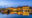 an image at dusk before it is completely dark of a row of yellow buildings along a river and in the water there is the reflection of lights all along and on the left hand side a wooden boat is parked in the water