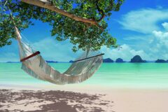 a white hammock hangs from a tree branch over a stretch of perfect white sand and in the background blue sea and in the distance a variety of limestone karsts come out of the water