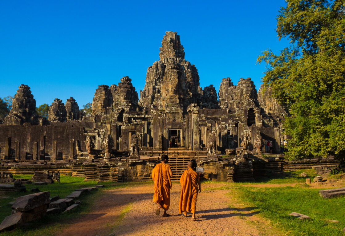 two monks in bright orange robes walk along a sand path towards the temple of angkor wat with it's different heighted towers and a bright blue sky is in the background