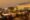 Upscale rooftop bar and lounge at dusk featuring Art Deco-inspired design with illuminated curved glass bar area, decorative brass brewing equipment centrepiece, wicker and wood outdoor furniture with orange and green striped cushions, wooden deck flooring, ornamental hedges, and warm golden ambient lighting overlooking a city skyline