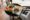 a person carries a black circular tray and on that are two rectangular plates, one with a salad and the other has a bowl of food and two pieces of bread on it and a wooden table in the backgrond