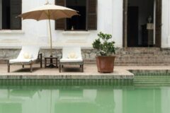 picture of the pool at Satri House taken from water level with sun loungers and umbrella and on the right some brick stairs lead to an open door and two windows with brown shutters go the left