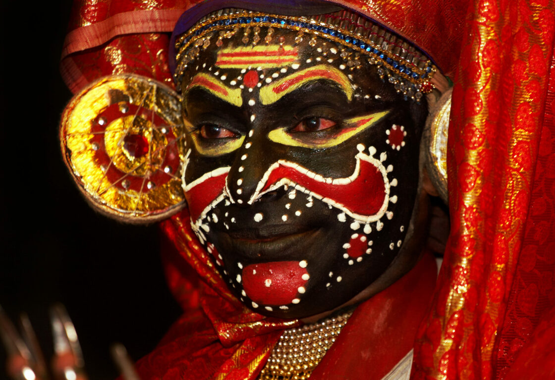 a close up of a persons face painted black with patterns on painted in white, red and yellow and they were a red head piece of a Kathakali dancer in india