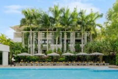 Viroth's Hotel in cambodia a white building with a pool and loungers stretching out in front and with tall palm trees growing in front