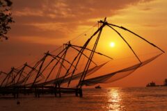 large fishing nets silhouetted against an orange sky as the sun sets, suspended over the water on wooden structures along the Cochin shoreline in india