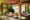 Adiwana Monkey Forest in Indonesia a wooden frame stands above a bar area lined with some bottles and chairs with thick white cushions line the bar and in the back ground plants and trees grow from flower beds