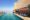 A blue and still pool runs along the side of a building overlooking a busy city in the background and along the edge of the pool are luxurious sun loungers with thick blue padding and curtains pulled to the side for privacy