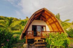 Komodo Resort a white hut with a wooden triangular roof and balcony with a set of wooden stairs and behind the hut a hit with palm trees growing