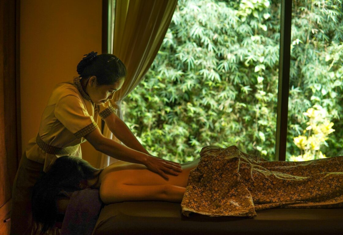 a woman lies face down on a bed with her body covered by a printed sheet and at her head another woman leans over giving a massage and a curtain reveals greenery in the background at Munduk Moding in Indonesia