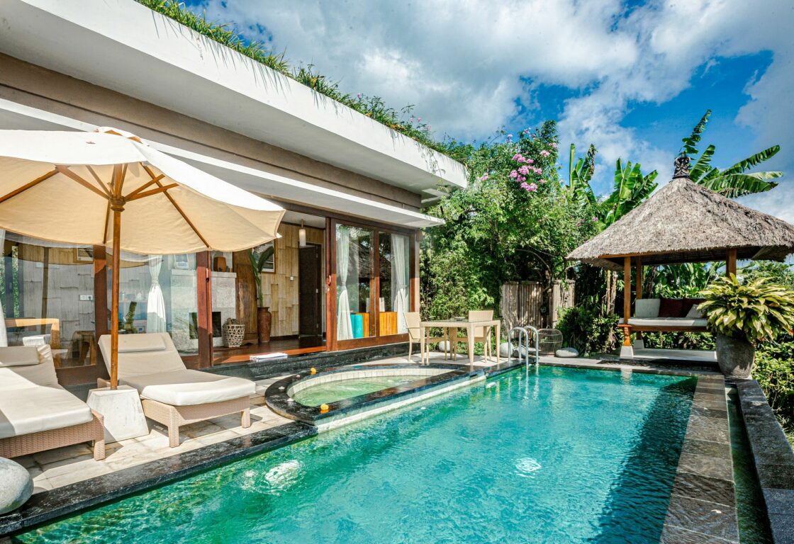 a beautiful pool with a covered sun lounger at the end in front of a one story bedroom with open doors and sun loungers with an umbrella at the edge of the pool at Munduk Moding in Indonesia