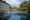 Plataran Heritage Borobudur in indonesia a large pool with blue sun loungers and umbrellas on the left hand side with a four story building behind that has doors with balconies and on the right hand side a stone wall with Hindu carvings with plants and grass growing along the top