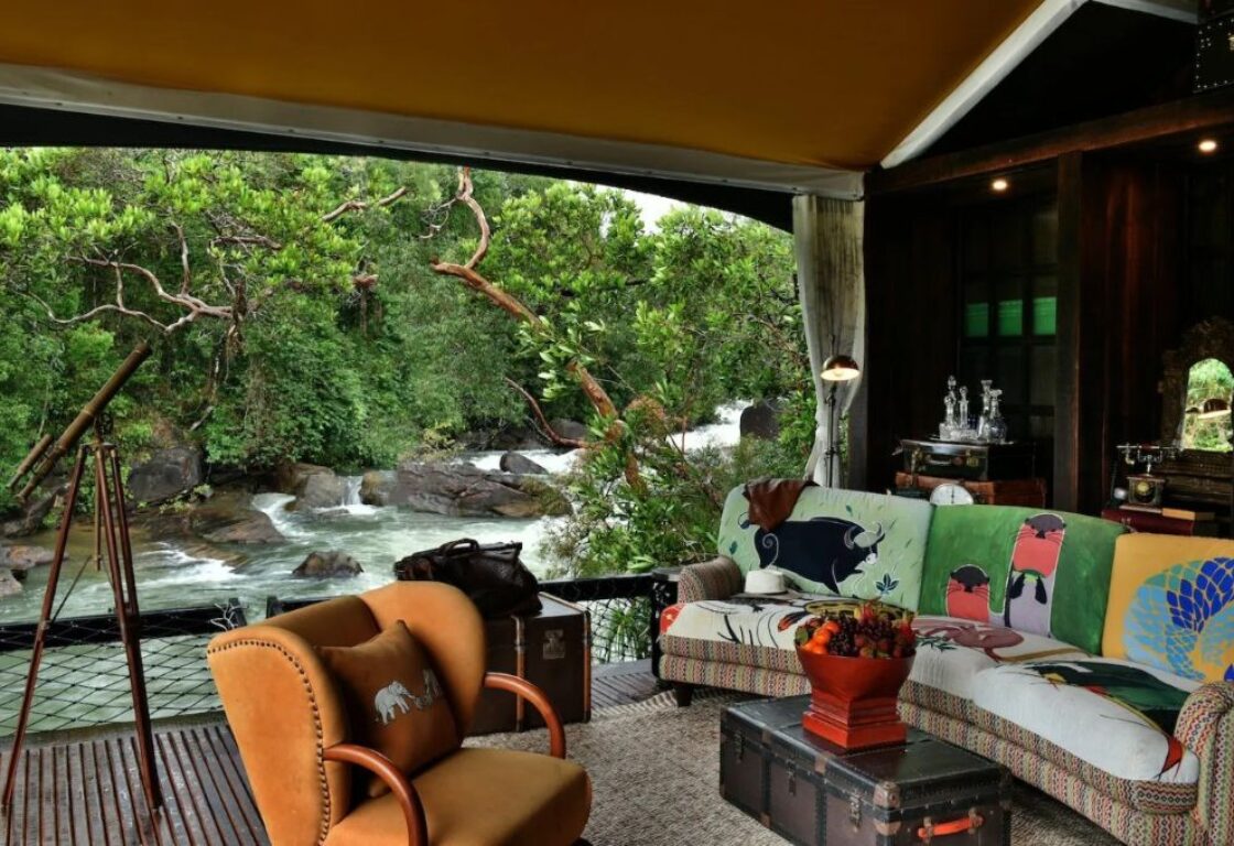 image of a cosy lounge with arm chair and sofa with colourful designs with an open view onto a river and trees at shinta mani wild in cambodia