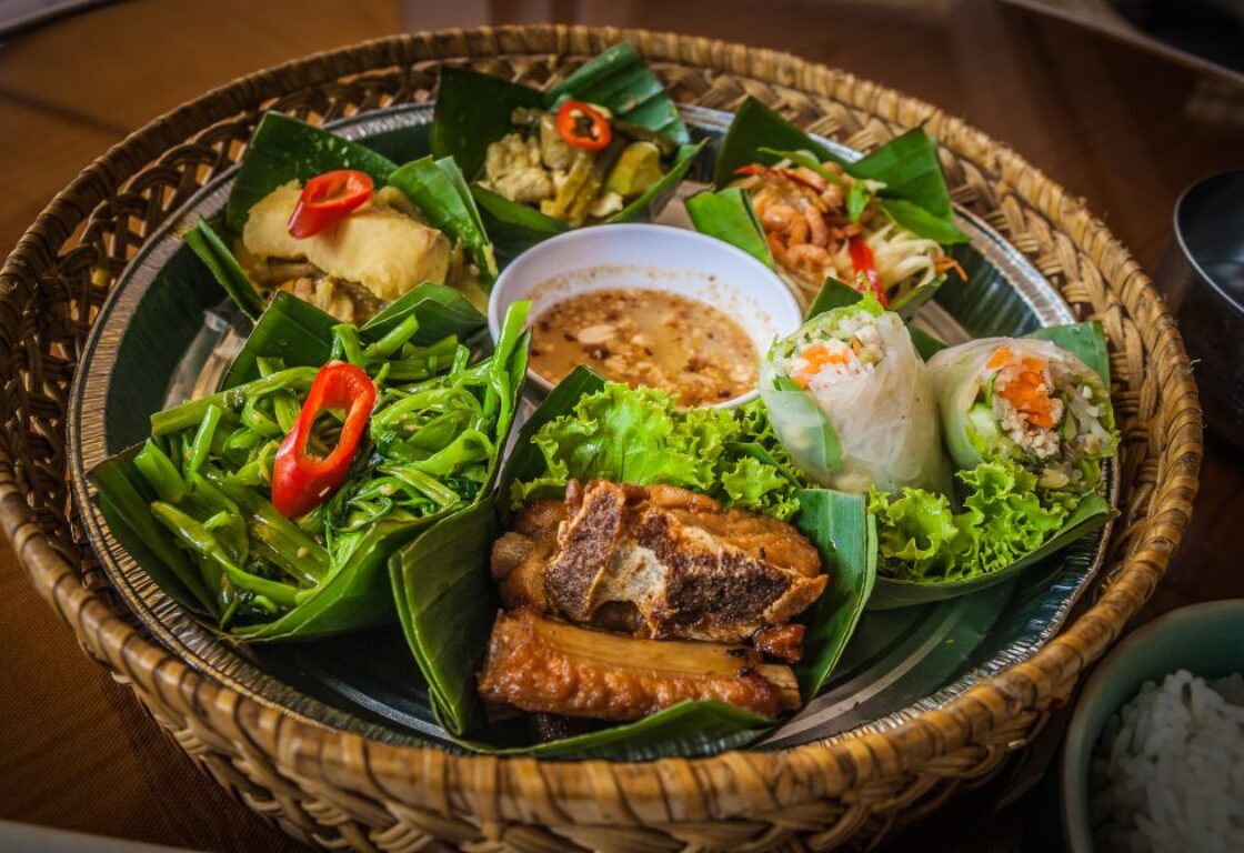 selection of different food on leaves served in a wooden basket including some meat, a large spring roll cut in half and in the middle a small pot with a brown sauce in