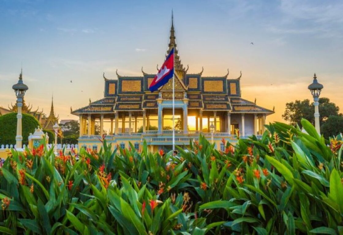 sunset behind gold roof silver pagoda with bushes in the front and cambodian flag in the middle