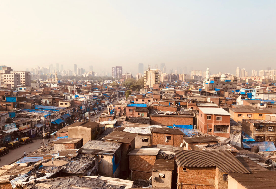 aerial view of lots of brown square buildings crammed into a street and on the other side of the road more brown buildings and some have blue sheets up and far in the distance a line of modern skyscrapers in mumbai india