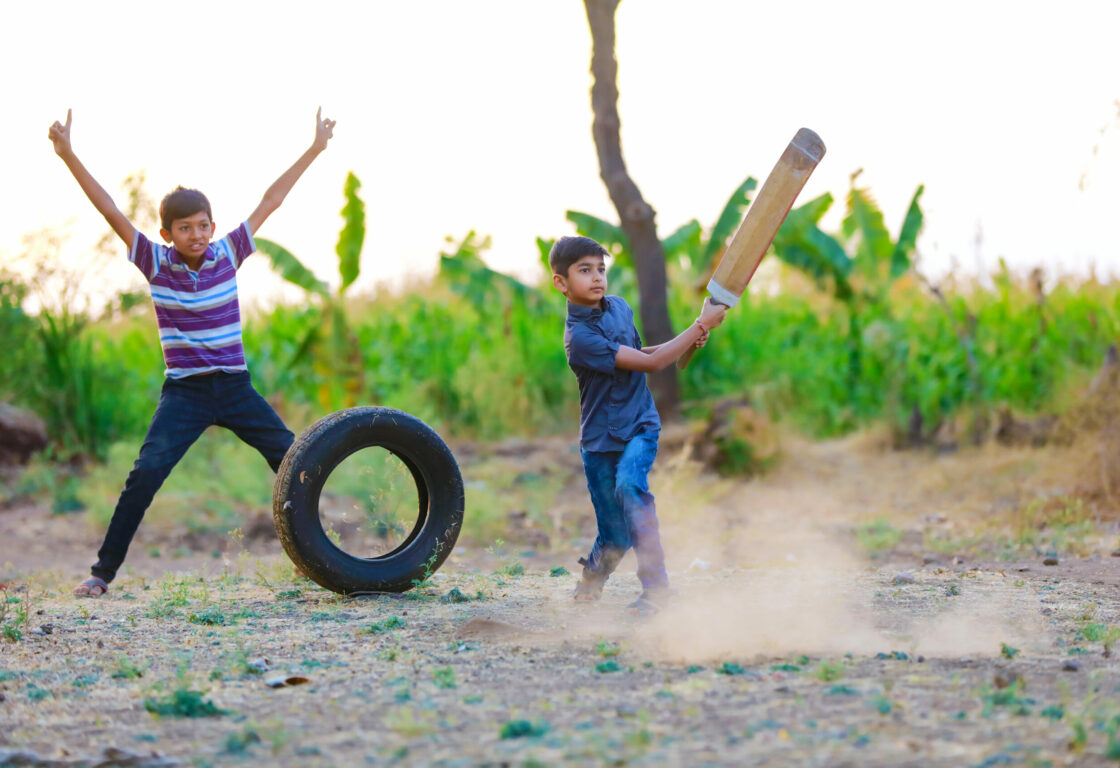a young boy swinging a cricket bat and another young boy with his arms above his head behind as they play cricket