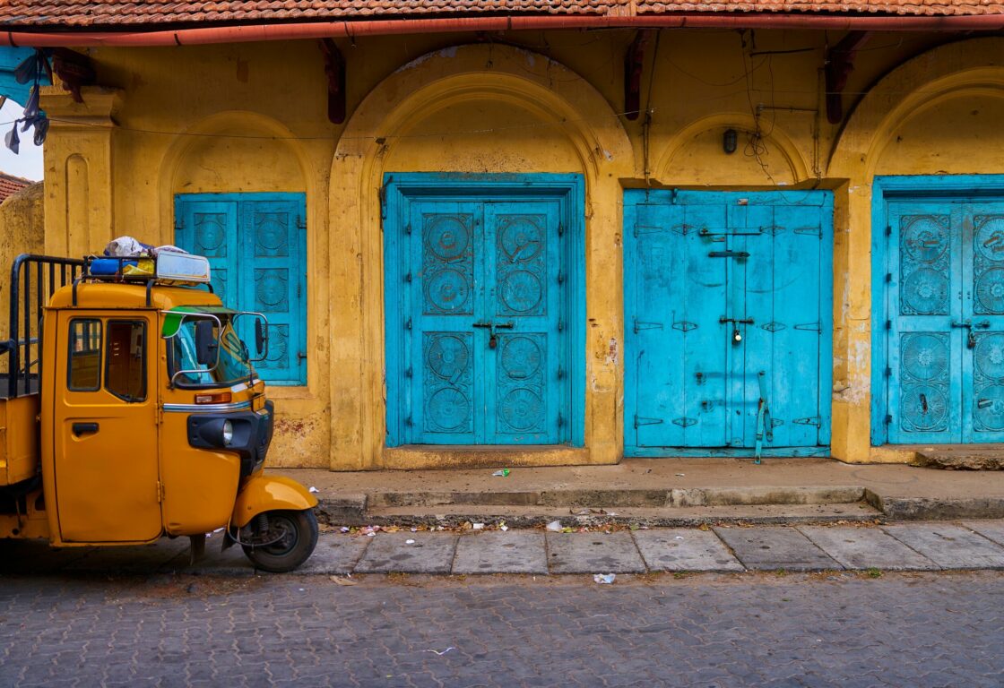a yellow wall with four large bright blue doors with carvings in and on the left the front of a yellow rickshaw in delhi is parked by the side of the road