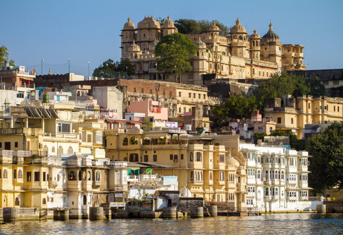 buildings up a river bank in udaipur, india, with a palace at the top of the hill