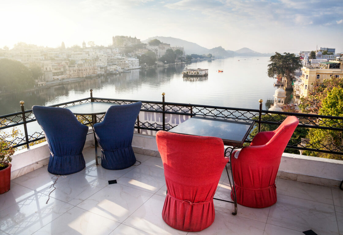 two tables with chairs on a roof terrace over looking a river in udaipur india, and buildings lining each side of the river