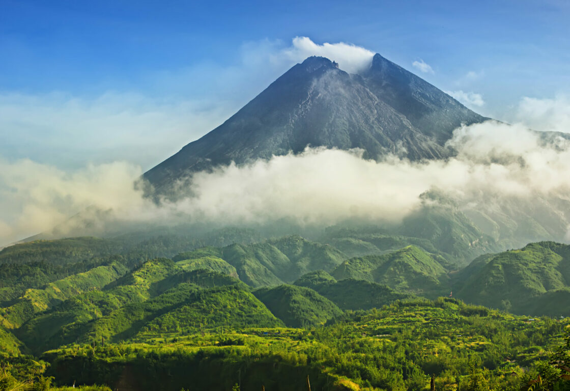 Mount Merapi in indonesia a tall volcano stands above smaller hills below it covered in grass and above those smaller hills a cloud hovers and a bright blue sky is in the background