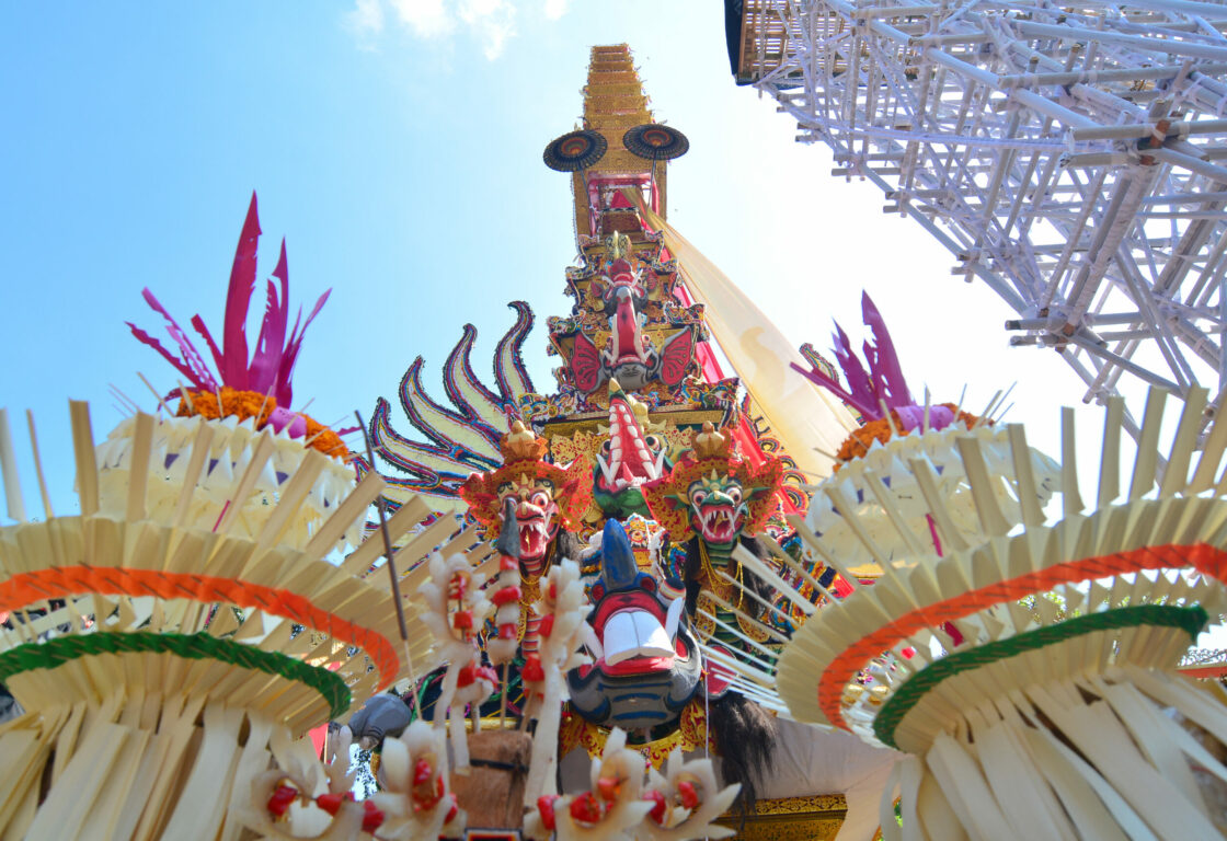 in the front of the image are what looks like upside down shuttlecocks and in the background a tall triangle goes in to the sky with bright colours and traditional balinese symbols including two small faces of dragons