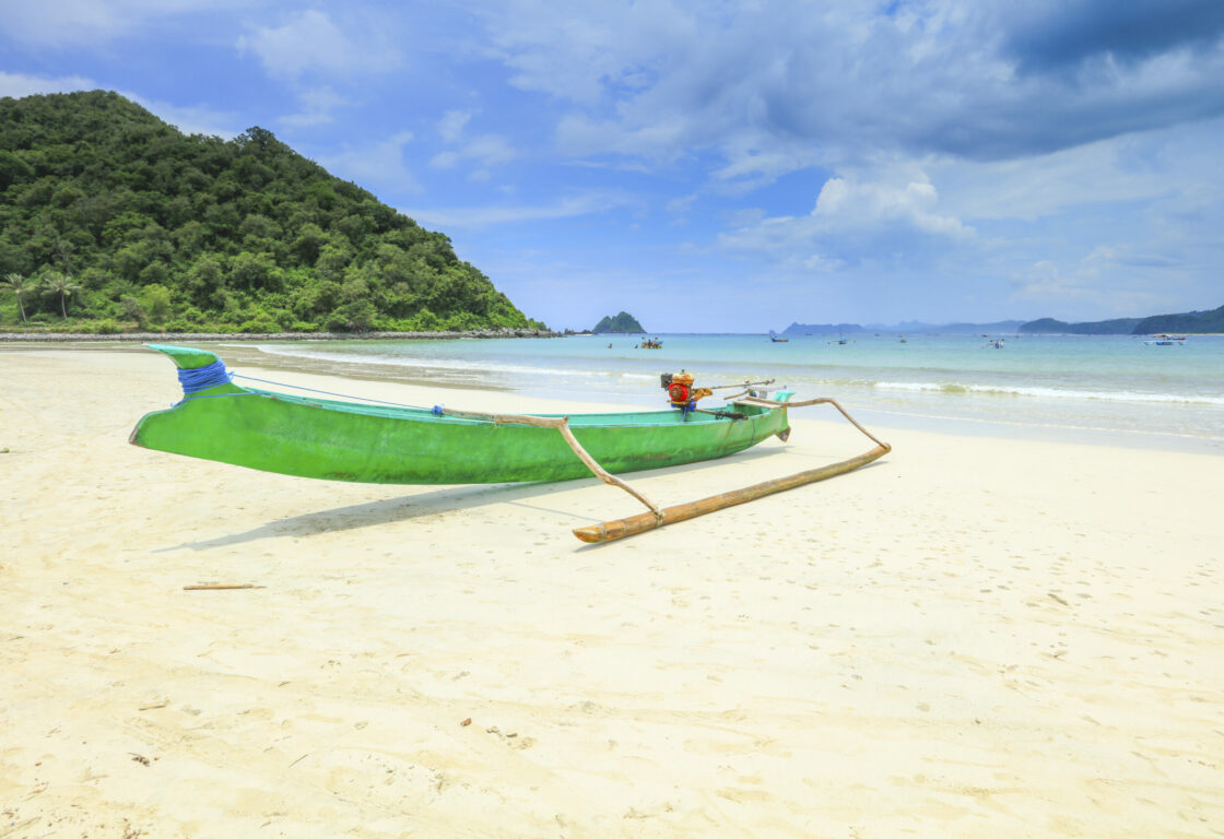 white sand on Senggigi with a green long boat on and in the back ground the sea has other little boats dotted around and on the left a hill covered with trees