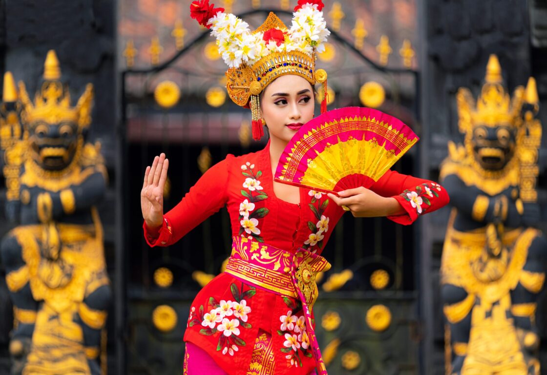A woman in a red dress with white flower details and a gold head piece with white flowers growing from the top holds a red fan partially in front of her face