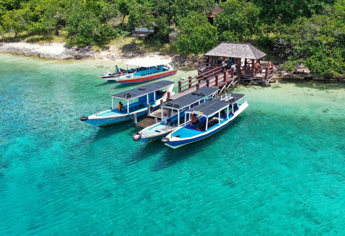 three boats parked next to a pier on Menjangan with beautiful crystal clear water and beyond the pier trees line the island and a small hut sits just along the line of trees