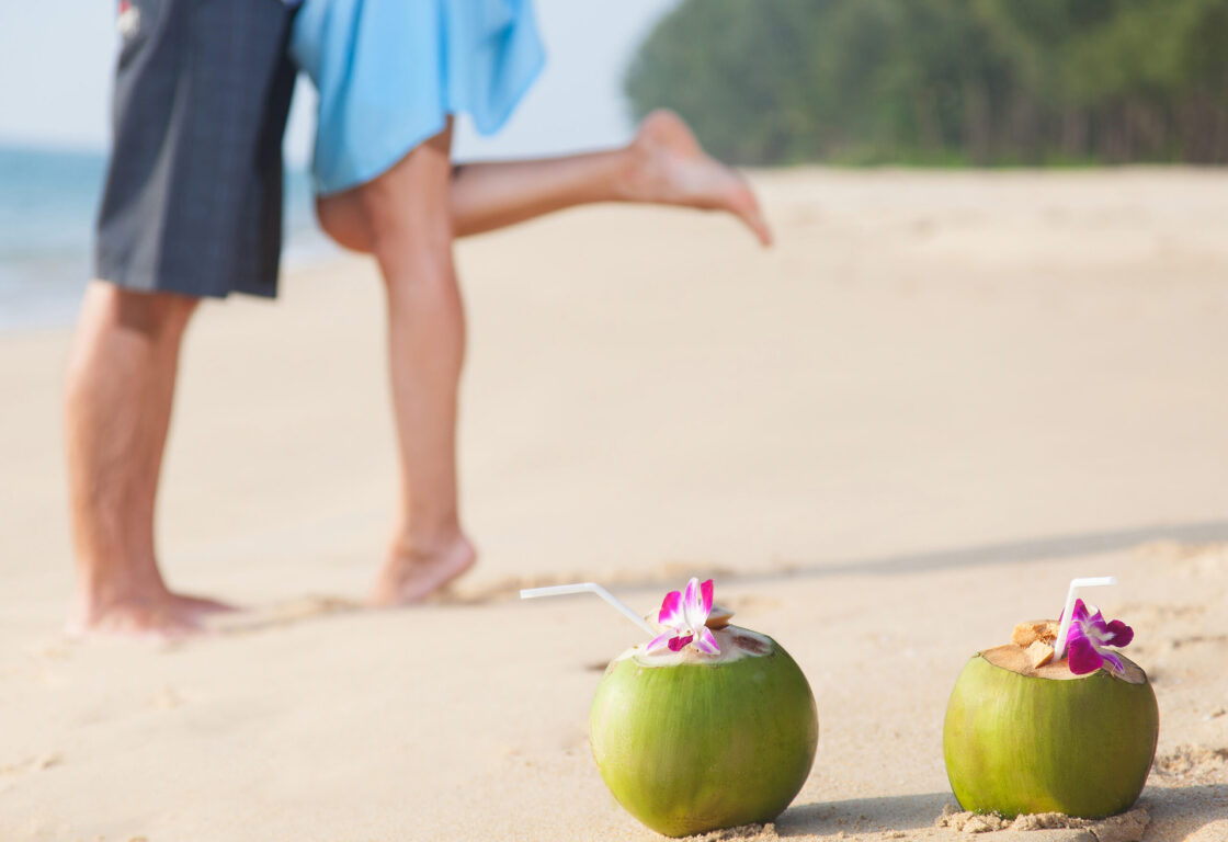 in the foreground of the picture are two green coconuts with a purple flower on top in the sands and behind are the legs of a man and a woman and she has one leg bent and raised at the knee
