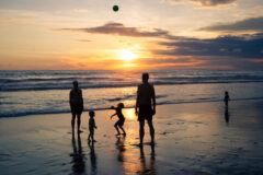 Children and adults throw a ball on the beach during sunset on the ocean background with floating surfers in bali
