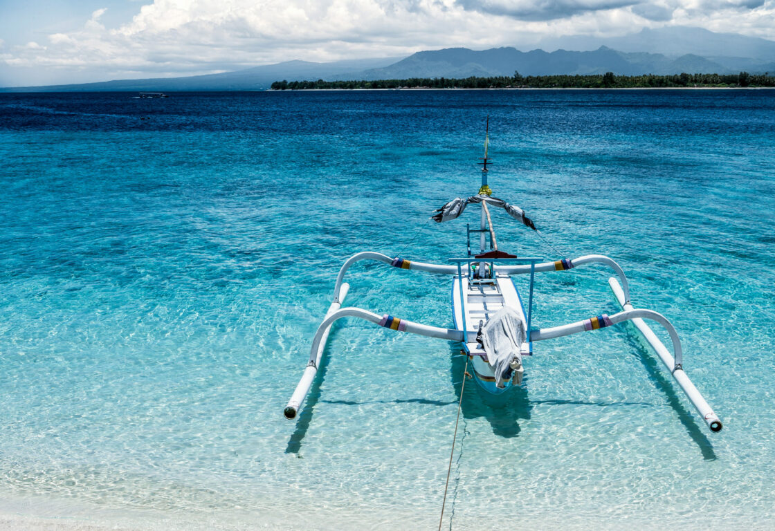 a white boat with two poles on either side to balance it sits on top of crystal clear sea in gili meno, indonesia