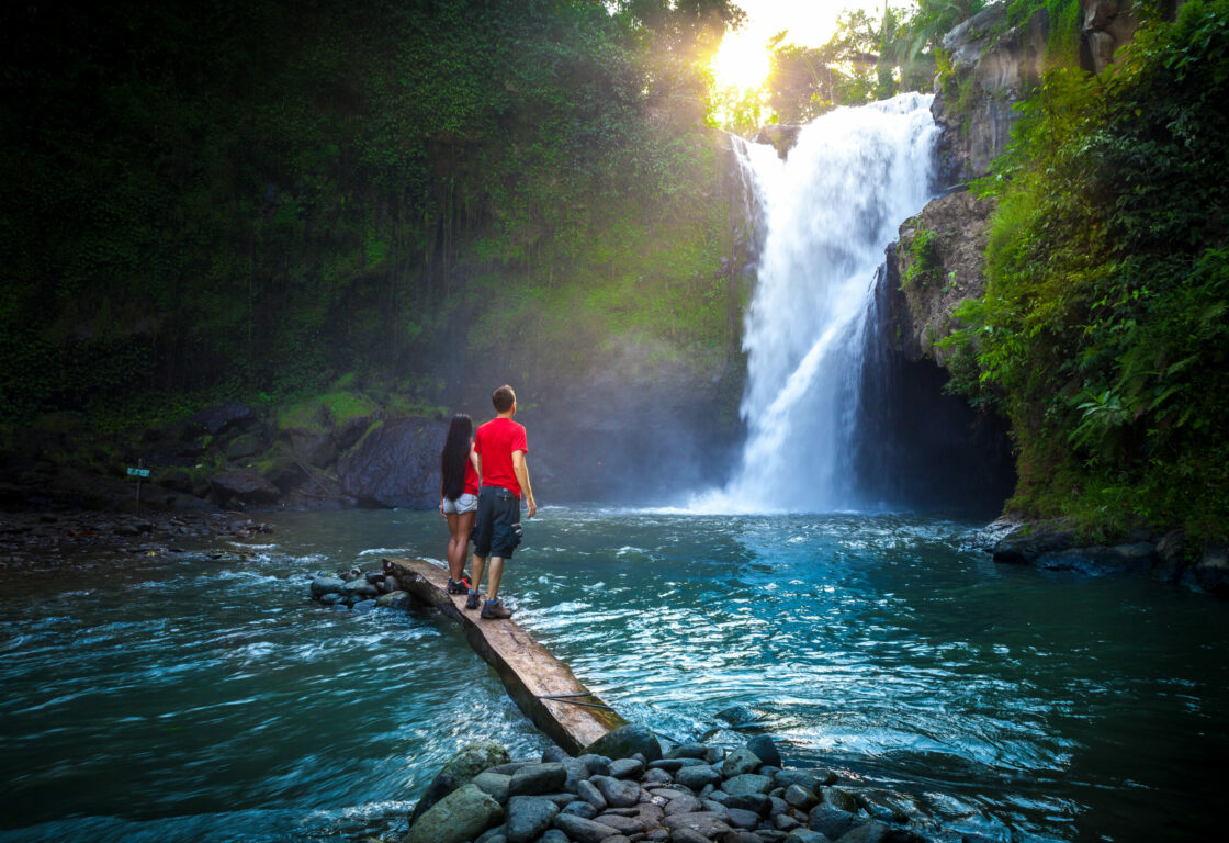 a man and a woman stand on a wooden plank that goes out onto a river and they look to a huge waterfall on the right hand side with lots of water rushing down