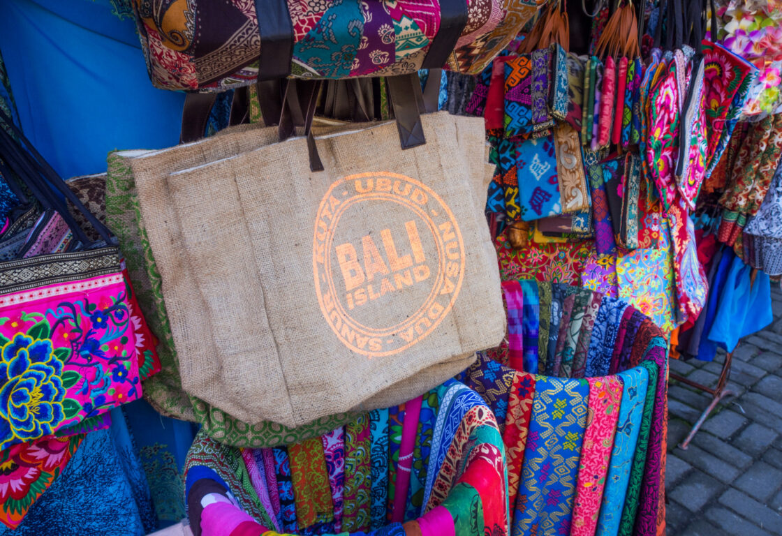 tote bags with bali island in a stamp in the middle of the bag hang in a market with lots of colourful bags and scarfs all around with different prints and patterns