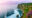 a stretch of cliff on the right hand side with lots of greenery growing and purple flowers growing in the corner and below there is the sea with white waves