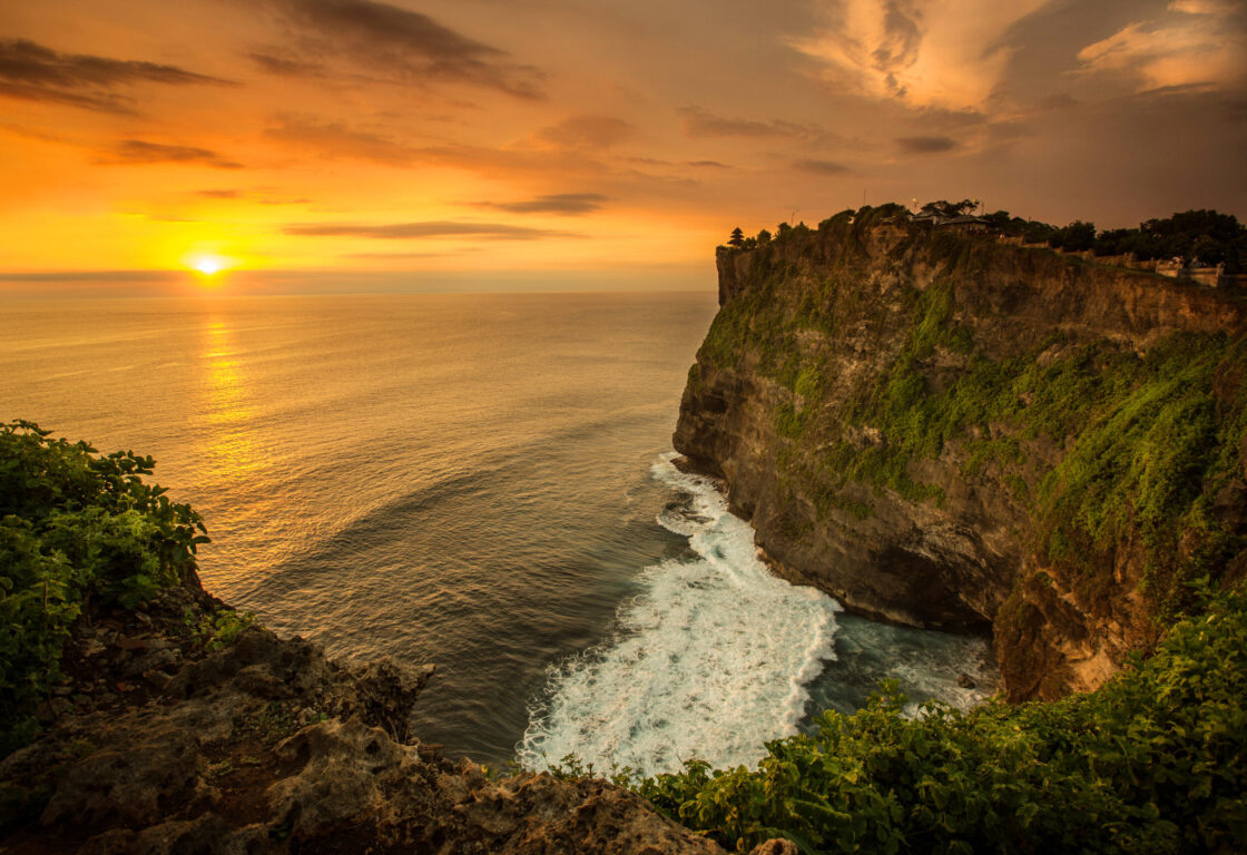 a section of cliff curves with the waves below and in the distance an orange sun sets in an orange sky and reflecting on the surface of the water