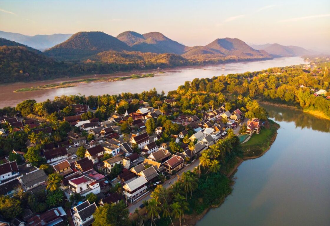 a river flows around a small village with the tops of houses in different shades of brown and in the background rolling hills stand tall above the water