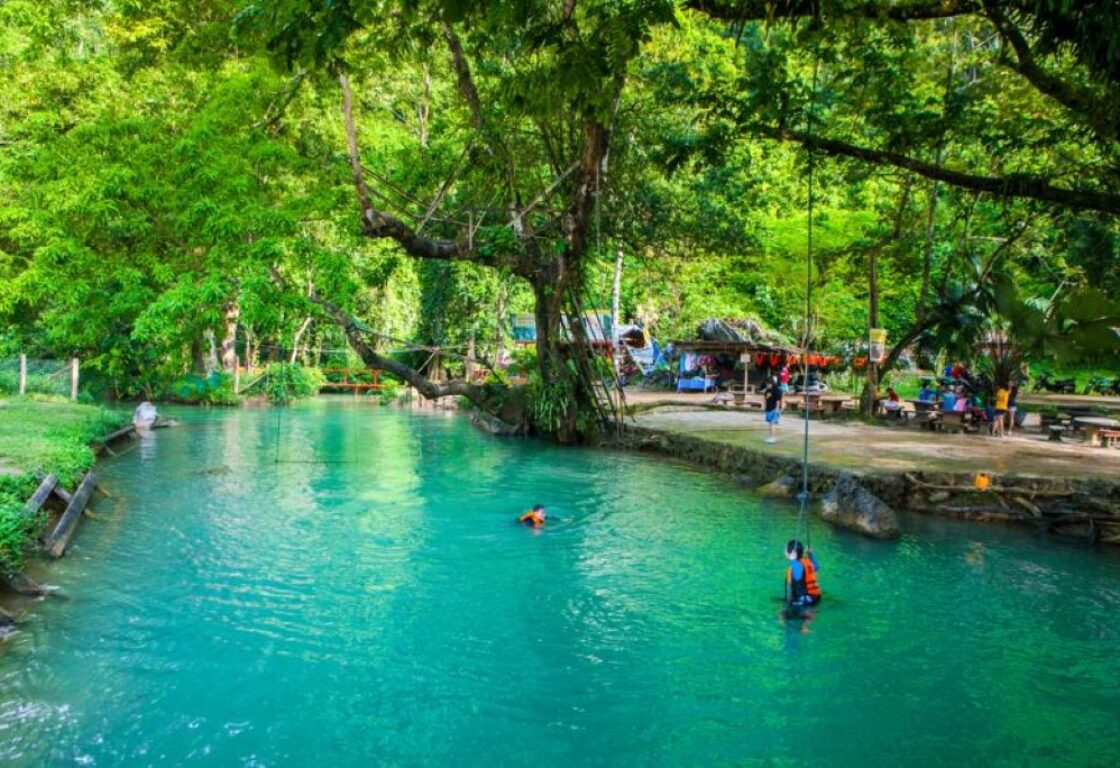 a large turquoise natural pool with two people swimming in it and trees frame the pool on one side and on the other a flat piece of either has some tables on