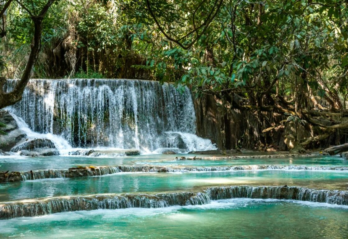 kuang si waterfalls in luang prabang, laos, with one tall level rushing water into a pool of turquoise blue water below that has a couple of different levels and trees surround the whole area