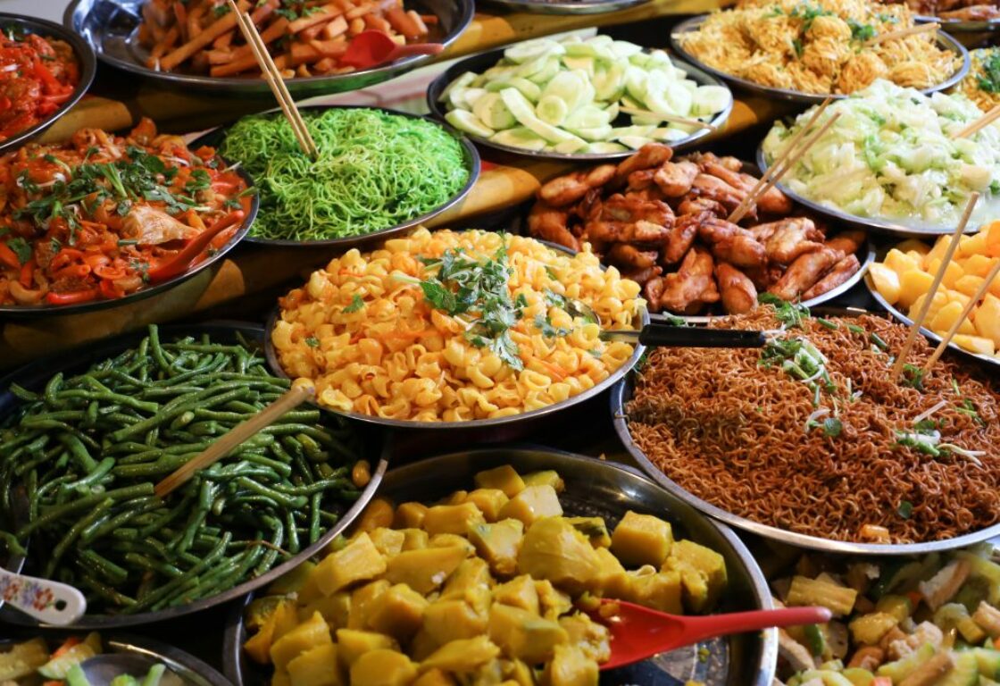 several metal bowls on display show a variety of food from vegetables, noodles, meats and a few have chopsticks in the food