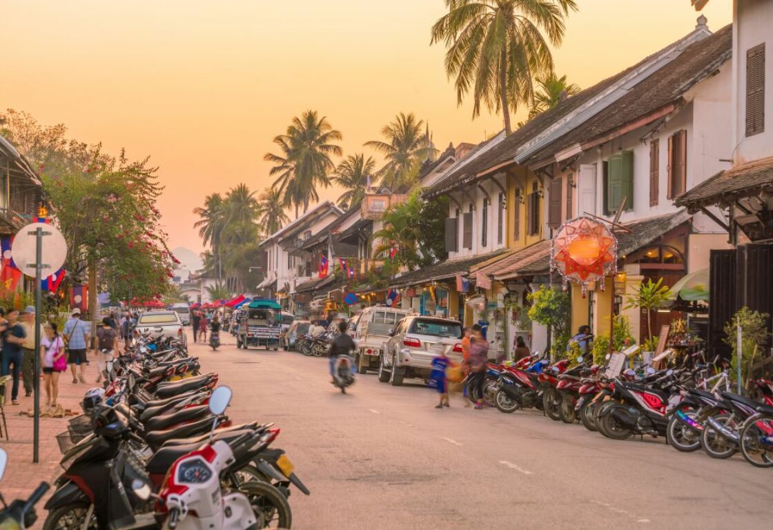 sunset over a busy street in Luang Prabang, Laos with mopeds lined all along the road and white houses with coloured shutters on one side