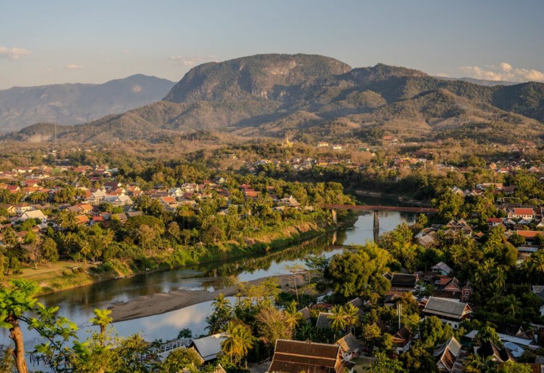 in the back ground a mountain stands tall and underneath that a river with a long bridge crossing connect two sides of trees and houses