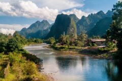 a large river runs down the middle of the picture with a bank on the left side filled with greenery and on the right mountains of different heights stand tall and reflect on the water