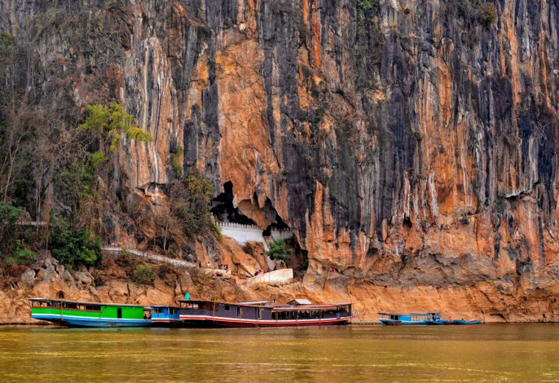 a tall cliff with jagged side and a small clearing reveals a white wall and traditional laos boats are parked at the foot of the cliff on a river in laos