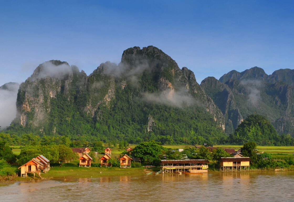 the jagged mountain tops against a blue sky with clouds and mist and at the bottom some fiends and trees next to a river and on the banks of the rive little wooden huts on stilts