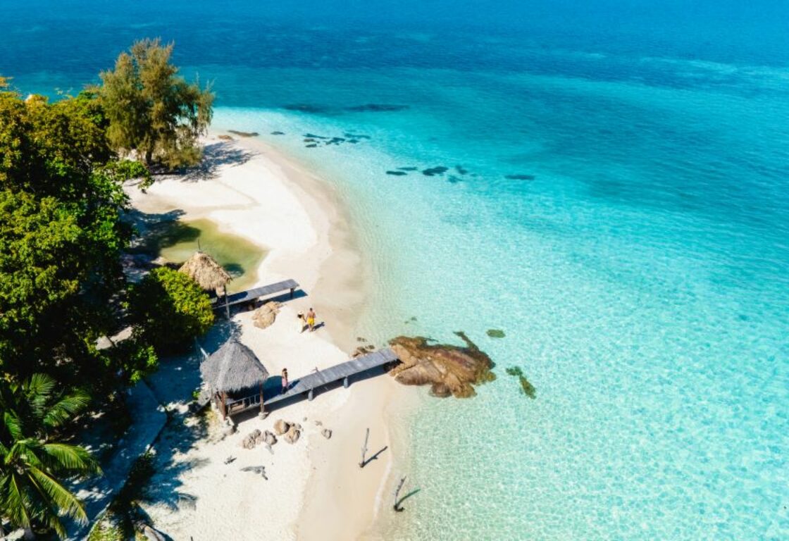 a small island with white sand and a small forest has a hut on and a pier that goes over the sand and leads into the sea that is bright and vibrant shades of blue