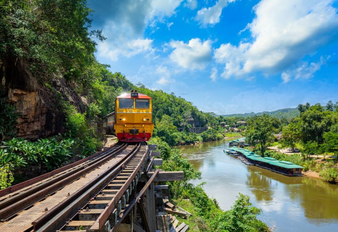 train on tracks at the edge of a hill in kanchanaburi with a yellow train coming along the track and a river below and green trees and countryside surround it all